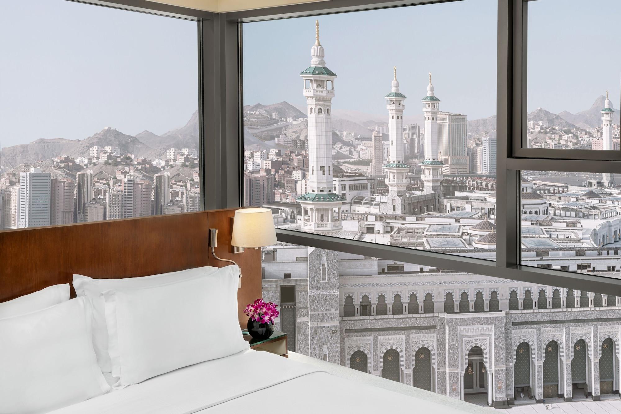 Hilton Suites Jabal Omar Makkah Mecca Exterior photo The photo depicts a hotel room with a modern design. In the foreground, there is a neatly made bed with white linens and a bedside lamp. A small flower arrangement adds a touch of color to the room. Through large windows, a stunning view of a citysca