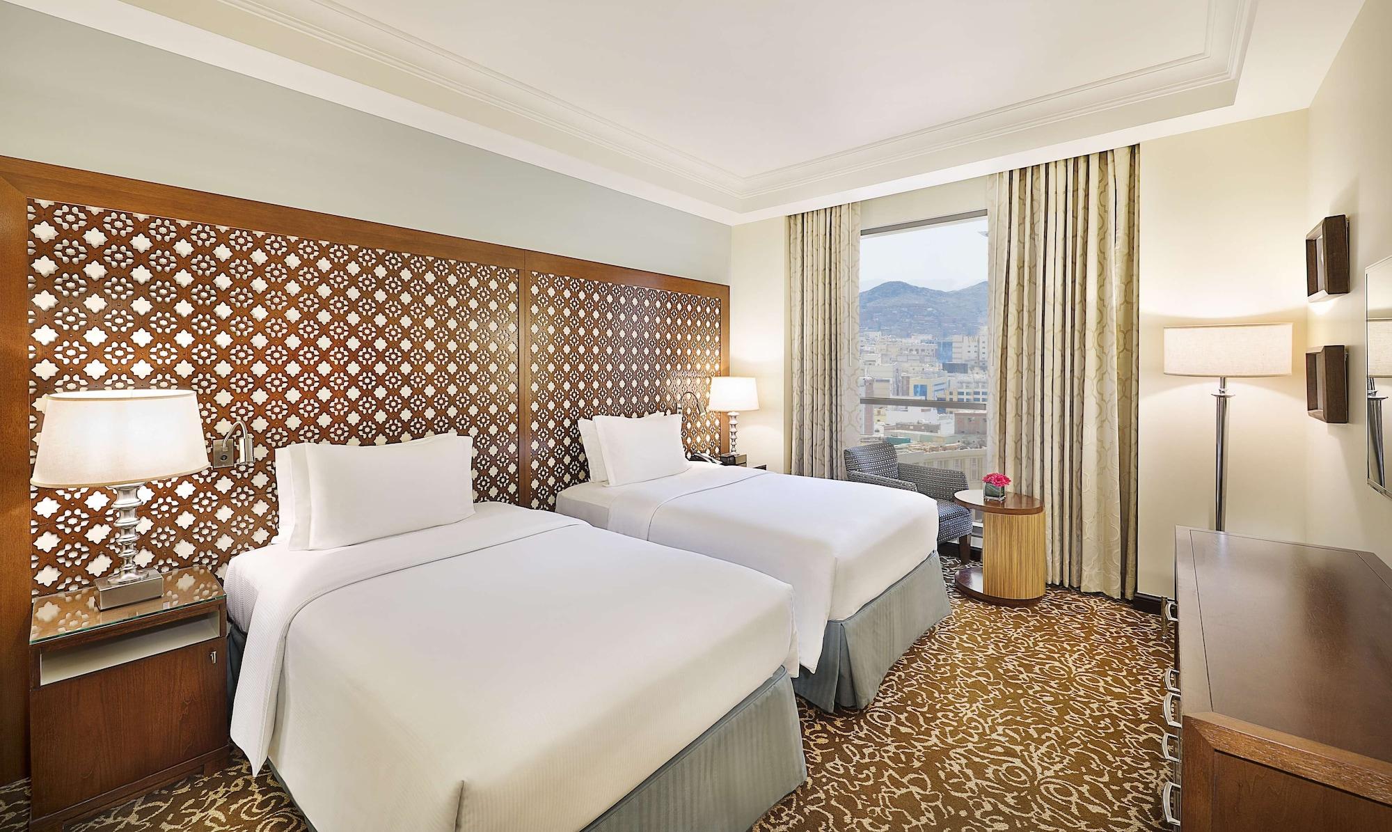 Hilton Suites Jabal Omar Makkah Mecca Exterior photo The photo shows a hotel room featuring two beds with white linens. The beds are positioned against a decorative wooden headboard with an intricate lattice design. There are bedside lamps on either side of the beds. A large window allows natural light