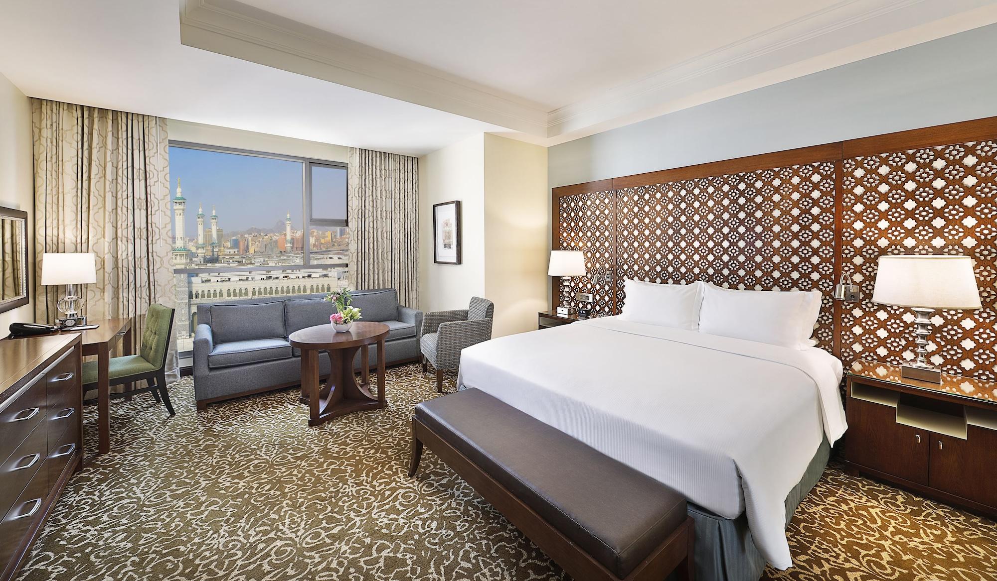 Hilton Suites Jabal Omar Makkah Mecca Exterior photo The photo shows a modern hotel room featuring a large bed with white linens positioned against an intricately designed wooden headboard. There are two armchairs and a small table in the corner, and a decorative vase with flowers on the table. A large