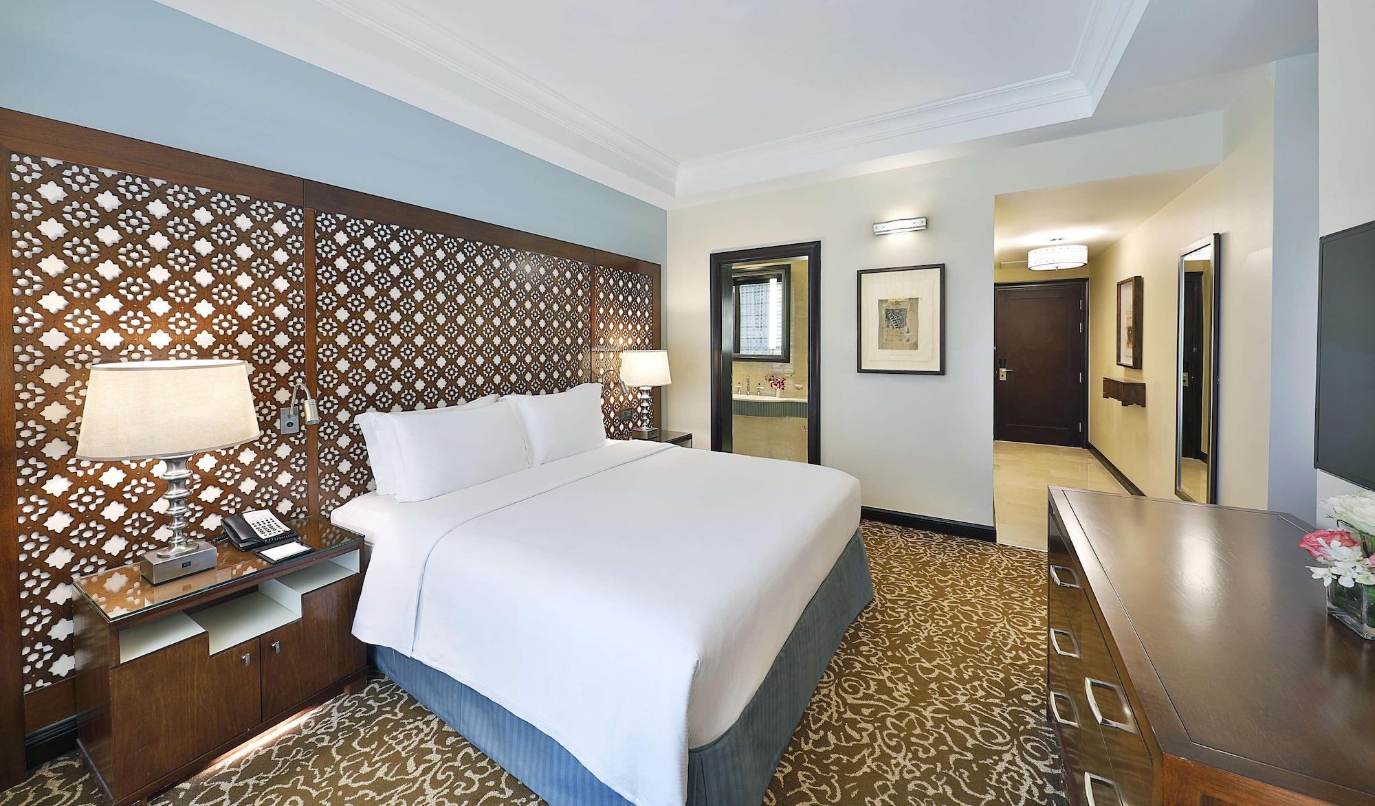 Hilton Suites Jabal Omar Makkah Mecca Exterior photo The photo shows a hotel room featuring a double bed with white linens. The headboard is made of decorative wooden panels. On either side of the bed, there are bedside tables with lamps. The room has a warm color scheme with a patterned carpet. In the