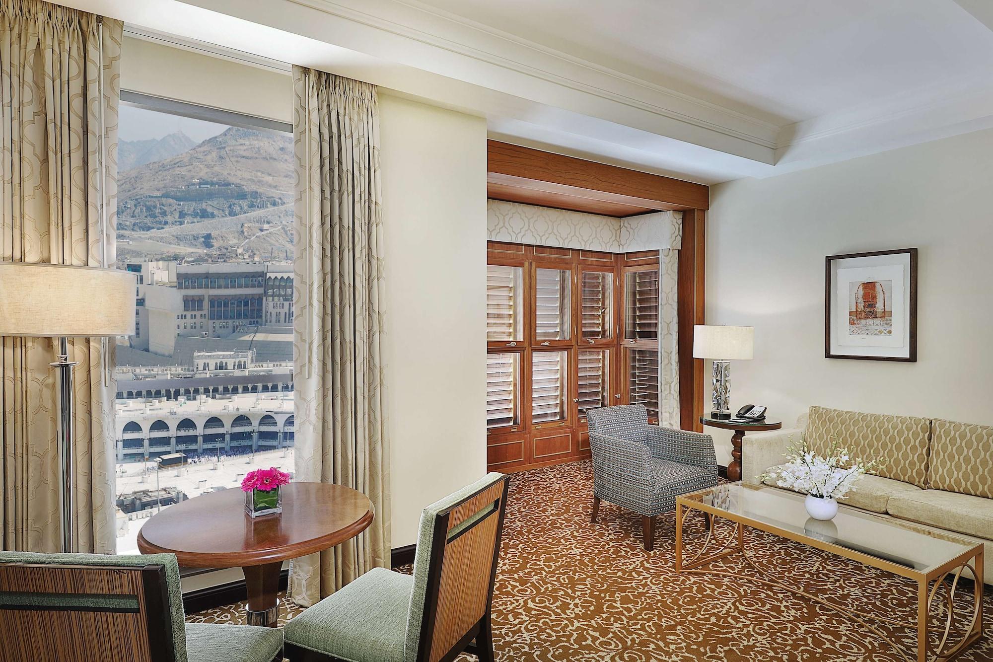 Hilton Suites Jabal Omar Makkah Mecca Exterior photo The photo shows a luxurious hotel room with a welcoming ambiance. There are large windows that provide a view of the outside, suggesting a well-lit space. The room features a seating area with a couch and chairs, alongside a small round dining table.