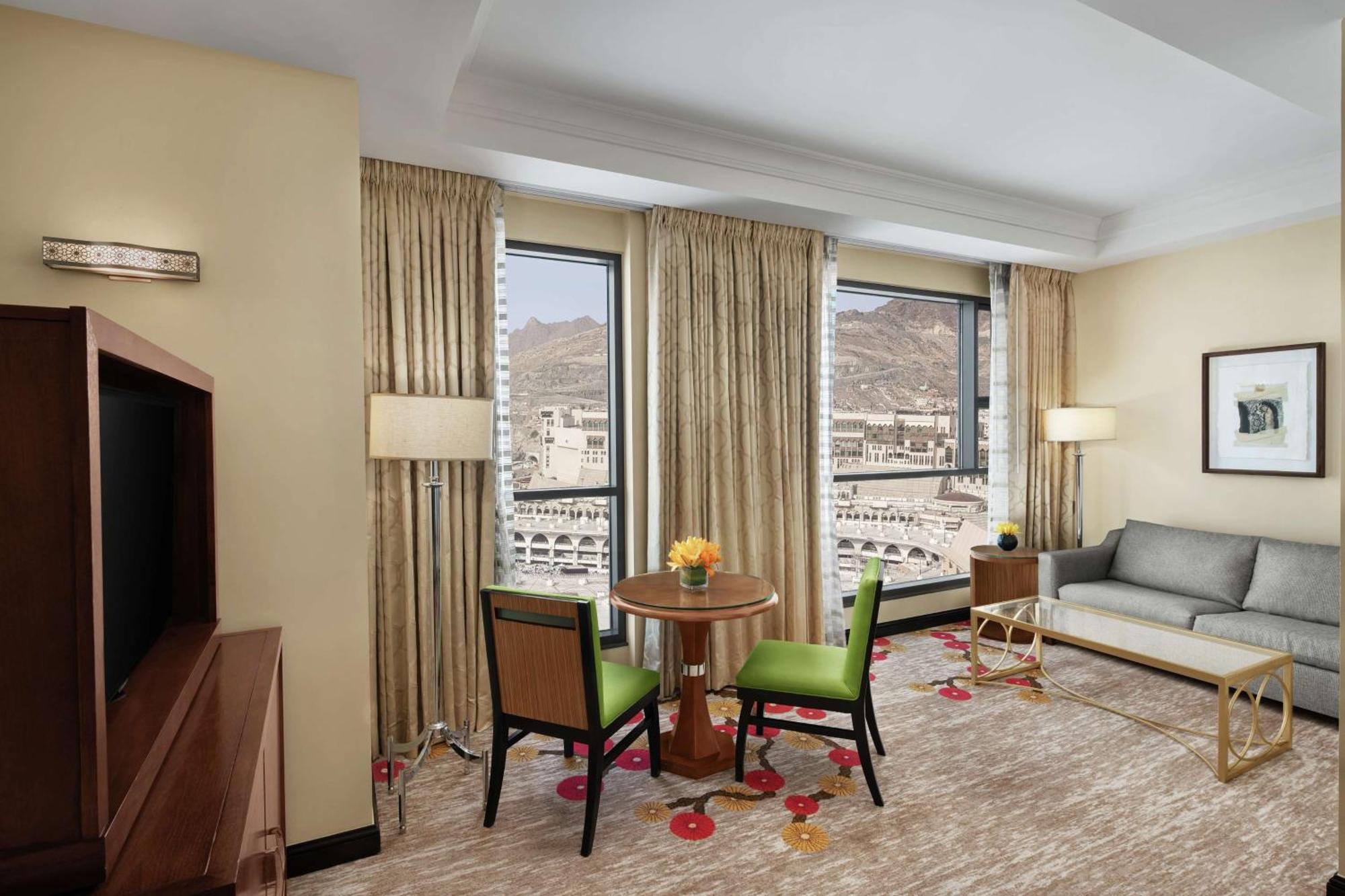 Hilton Suites Jabal Omar Makkah Mecca Exterior photo The photo shows a hotel room interior featuring a cozy sitting area. There is a small round dining table with two green chairs, and a vase of flowers sits on the table. A comfortable gray sofa is placed against one wall. The windows are adorned with 