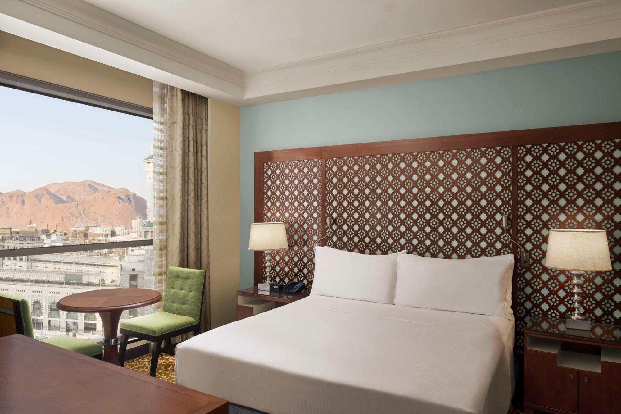 Hilton Suites Jabal Omar Makkah Mecca Exterior photo The photo shows a hotel room featuring a large bed with white linens and a decorative headboard. Beside the bed, there are two lamps on nightstands. A small round table with a green chair is positioned near the window, which offers a view of a mounta