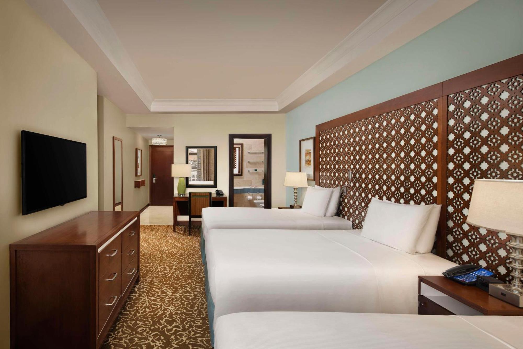 Hilton Suites Jabal Omar Makkah Mecca Exterior photo The photo shows a hotel room featuring two beds with white linens. There is a decorative headboard behind each bed, made of wood with a patterned design. The room has a warm beige carpet and light blue walls, giving it a serene atmosphere. A wooden d