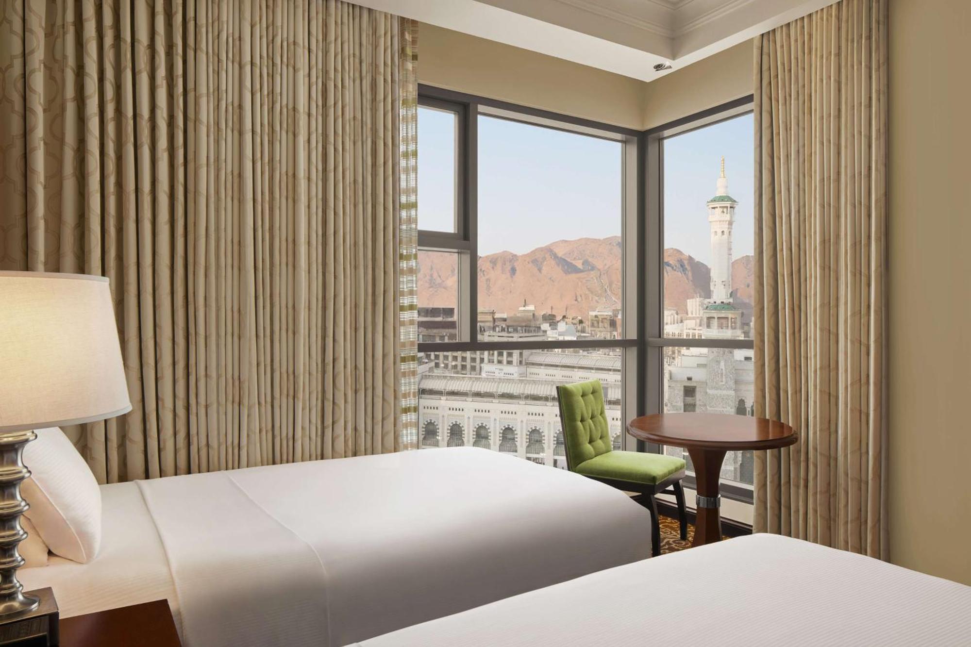 Hilton Suites Jabal Omar Makkah Mecca Exterior photo The photo displays a hotel room featuring two beds with white linens. Large windows adorned with curtain panels offer a view of a mountainous landscape outside. A small round table with a green chair is situated near the windows, creating a cozy area