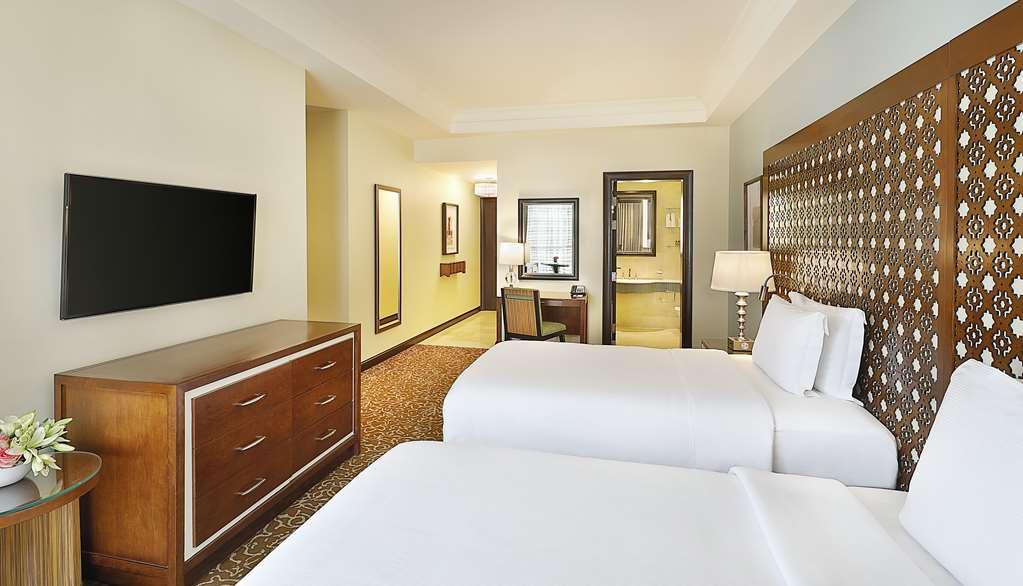 Hilton Suites Jabal Omar Makkah Mecca Room photo The photo shows a hotel room featuring two single beds with white bedding. There's a modern wooden dresser and a flat-screen television mounted on the wall opposite the beds. A small desk and chair are positioned near a window, and a bathroom is visi