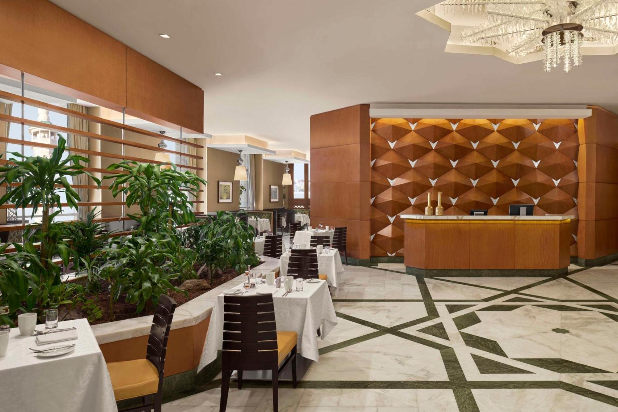 Hilton Suites Jabal Omar Makkah Mecca Exterior photo The photo depicts an elegant restaurant interior. The space features a sleek design with wooden elements and a stylish reception area. To the left, there are neatly arranged dining tables with white tablecloths, each set with cutlery and decorative t