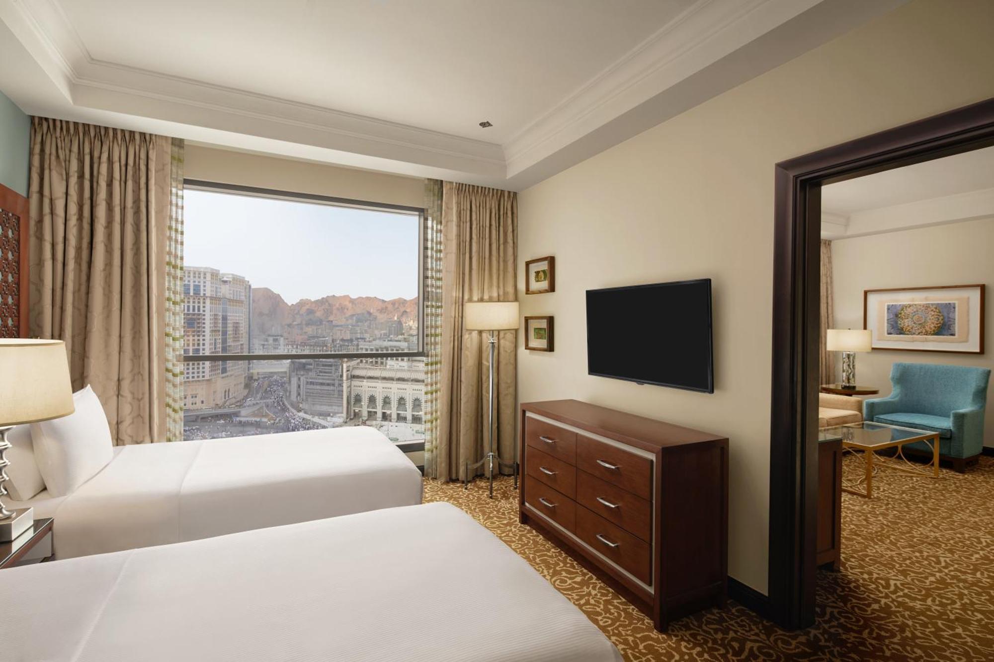 Hilton Suites Jabal Omar Makkah Mecca Exterior photo The photo shows a hotel room featuring two beds with white bedding. There is a large window with curtains that allow for natural light, offering a view of the outside scenery, which includes mountains. In the room, there is a wooden dresser and a fla