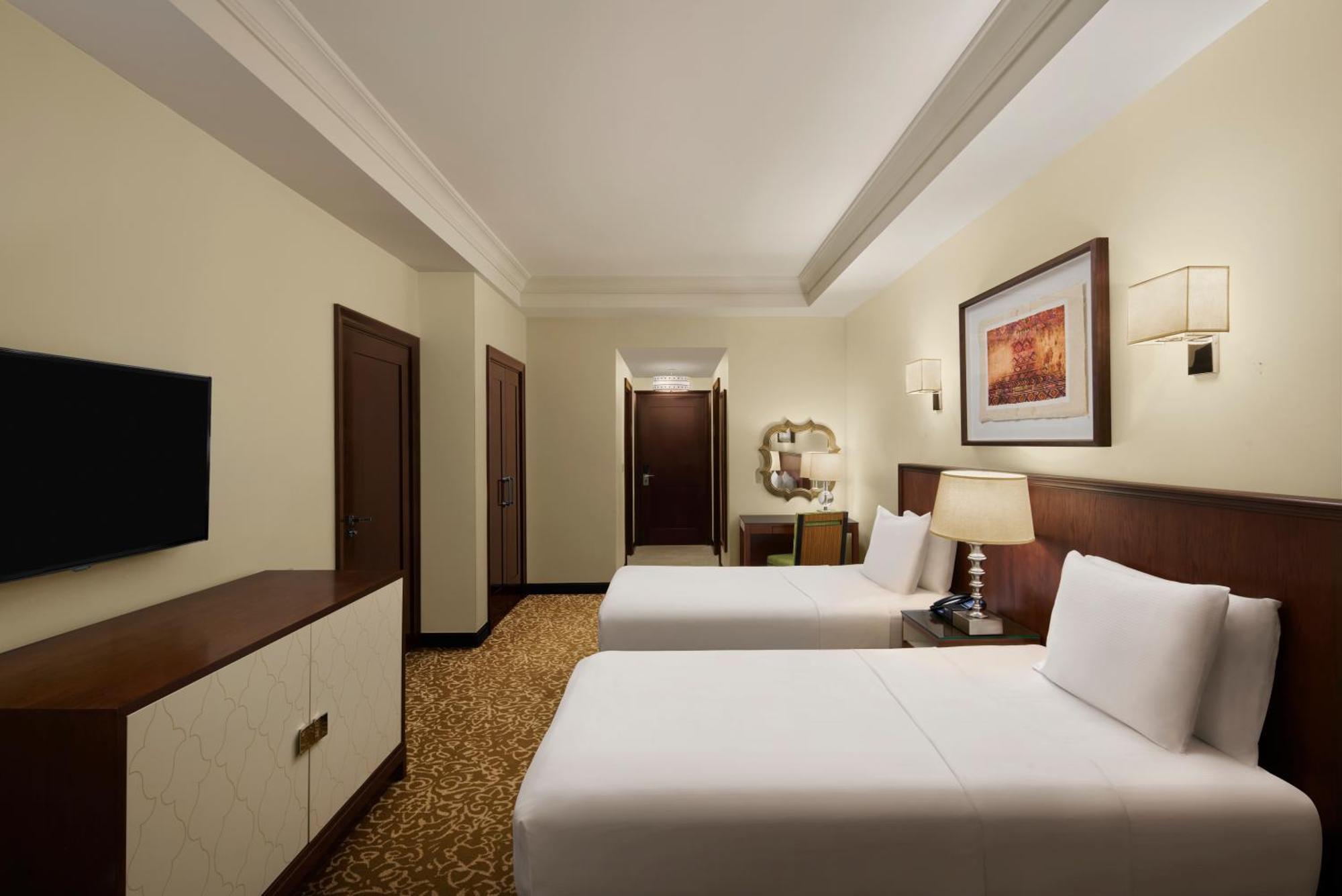 Hilton Suites Jabal Omar Makkah Mecca Exterior photo The photo shows a hotel room that features two beds with white linens. The room has light-colored walls and a warm-colored carpet. There is a wooden dresser with a flat-screen TV mounted on the wall opposite the beds. To the side, there are two doors