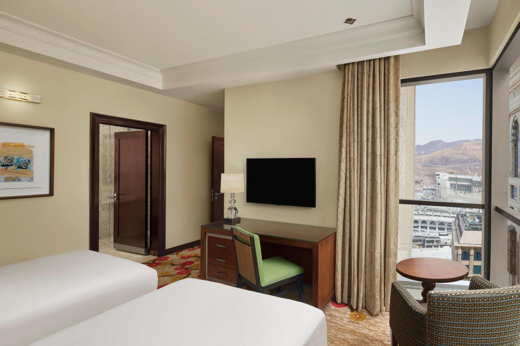 Hilton Suites Jabal Omar Makkah Mecca Exterior photo The photo shows a spacious hotel room featuring two beds, a desk with a green chair, and a flat-screen TV mounted on the wall. There's a large window with curtains that lets in natural light and offers a view of the outside. The decor is modern and w