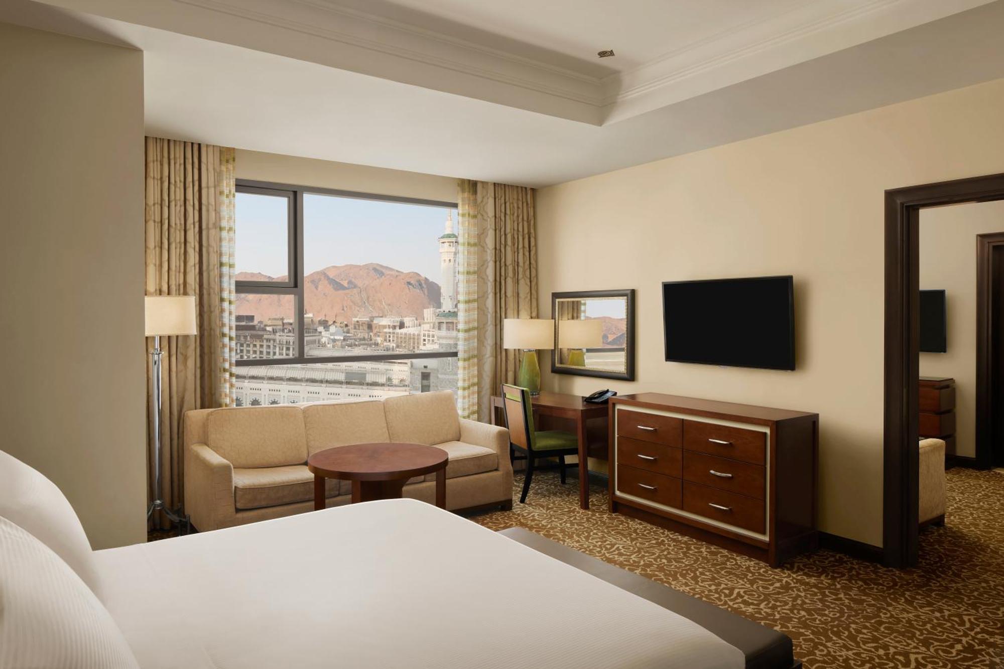 Hilton Suites Jabal Omar Makkah Mecca Exterior photo The photo shows a hotel room with modern decor. In the foreground, there is a large bed with white linens. To the left, a comfortable seating area features a sofa, a round coffee table, and a chair. A large window offers a view, likely of a mountain 