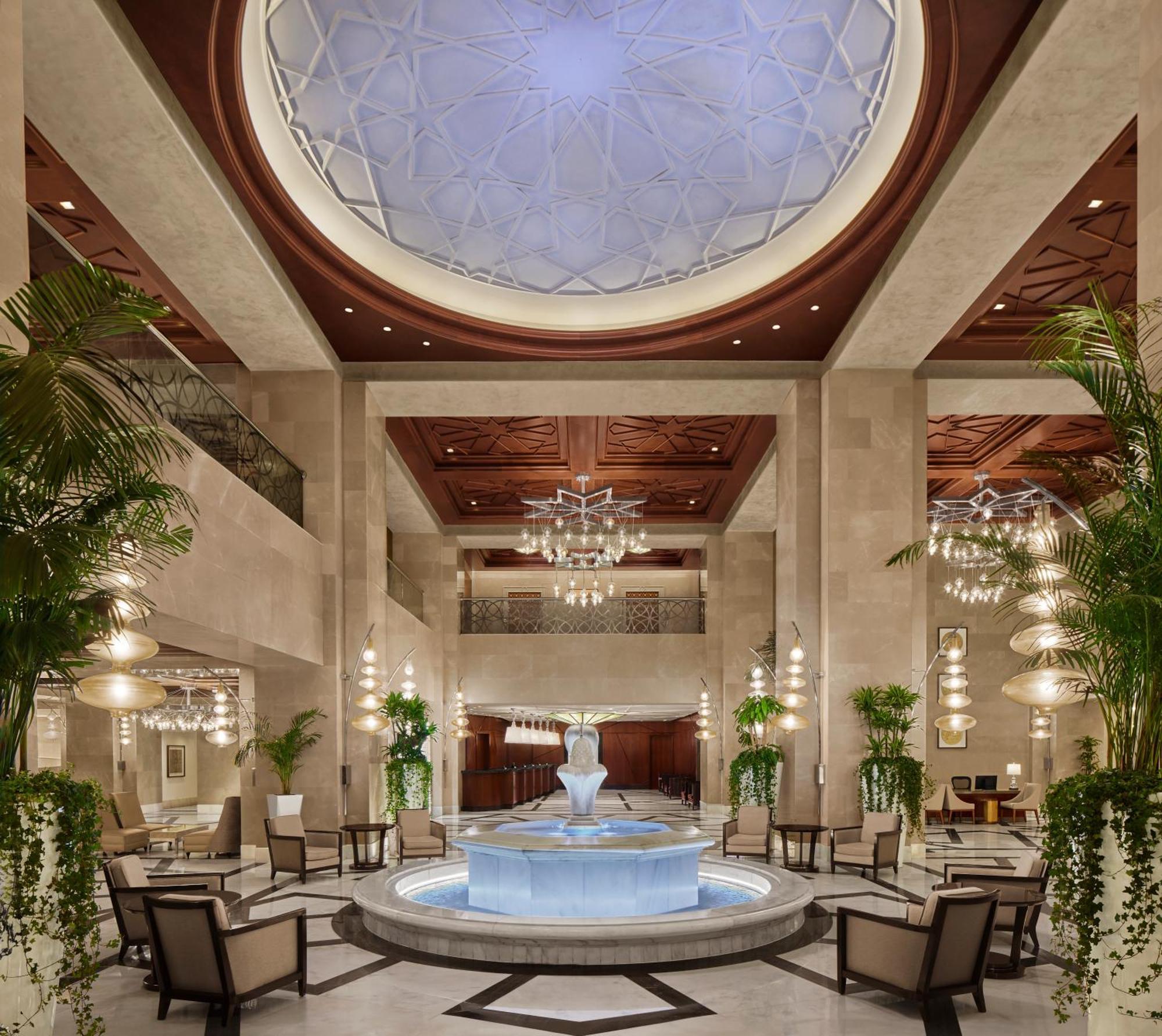 Hilton Suites Jabal Omar Makkah Mecca Exterior photo The photo depicts an elegant hotel lobby featuring a central water fountain. The ceiling has a decorative design, and there are large chandeliers hanging from it. The walls are adorned with light-colored stone, and lush green plants are positioned ar